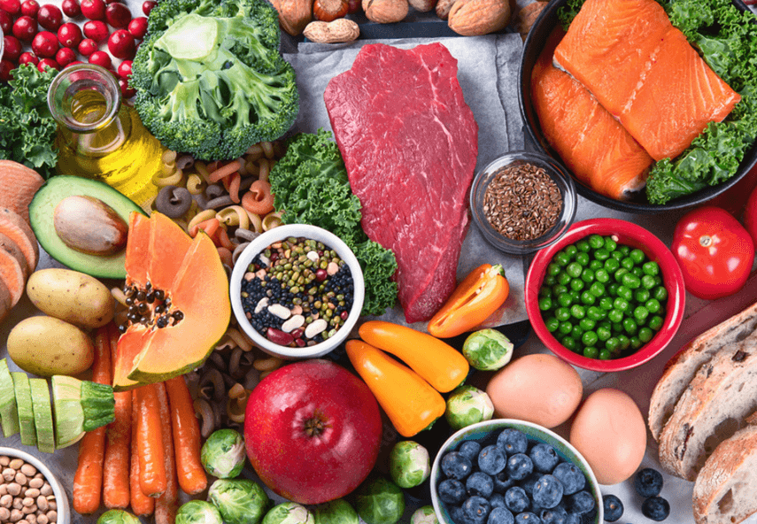 Meyveler, sebzeler, kuruyemişler, tohumlar ve etler dahil olmak üzere çeşitli sağlıklı yiyecekler bir masaya dizilmiş.