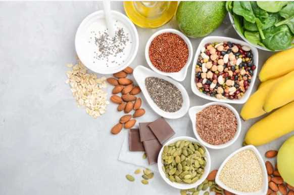 Tohumlar, tahıllar, kuruyemişler, baklagiller, muz, ıspanak, avokado, yoğurt ve bitter çikolata gibi çeşitli sağlıklı yiyecekler açık renkli bir yüzey üzerine dizilmiş.