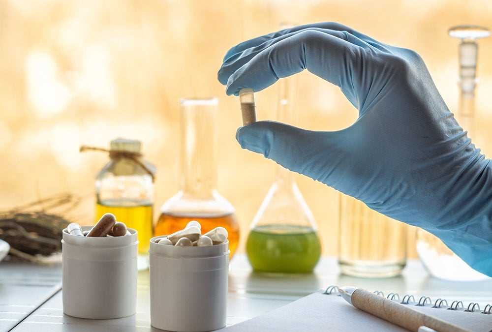 Pharma |Eine behandschuhte Hand hält eine Kapsel, während zwei Behälter voller Kapseln auf einem Tisch mit Laborglas stehen.