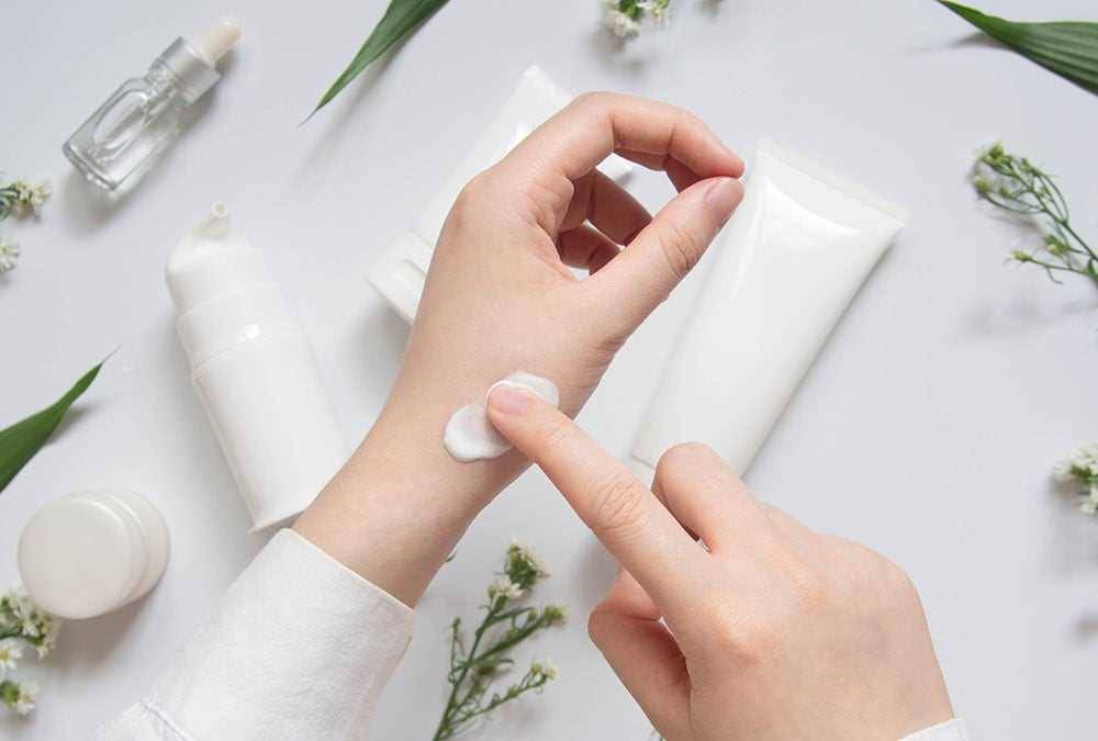 A hand is applying moisturizer on the back of a hand with 3 tubes of containers of cream in the background.