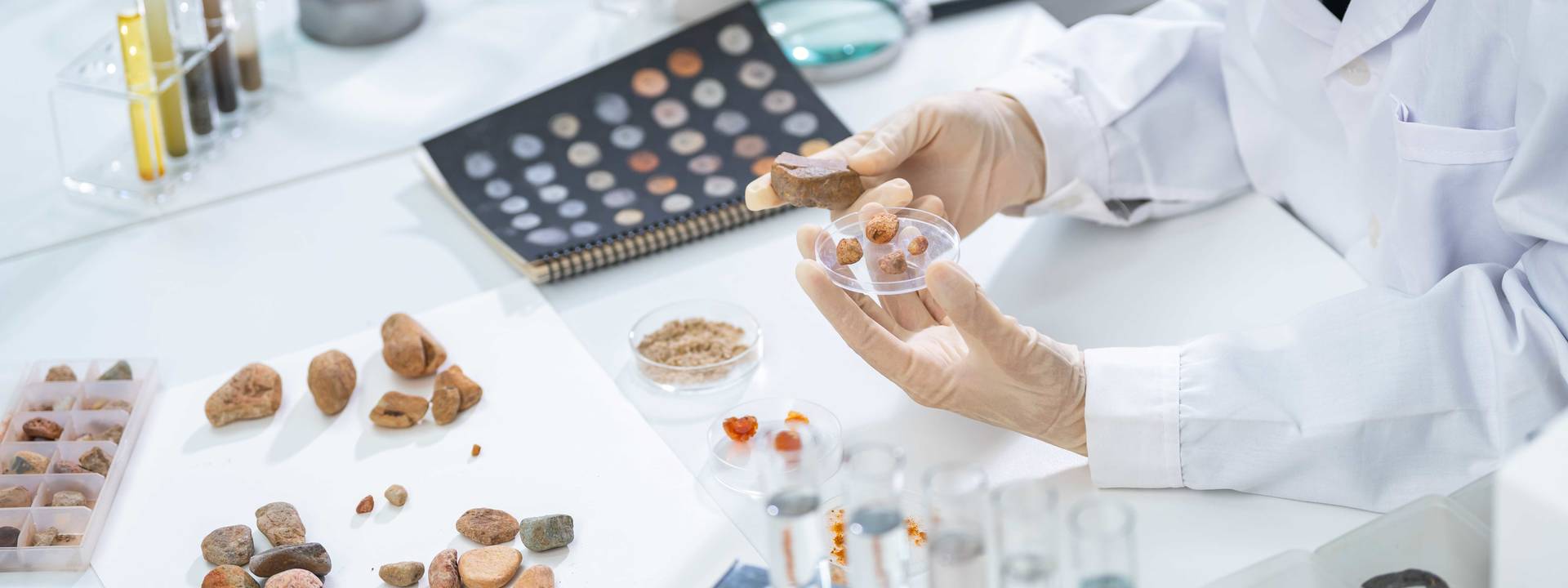 Pessoa com bata de laboratório e luvas a analisar amostras de rochas e sedimentos sobre uma bancada, com frascos, tubos de ensaio e um catálogo de minerais ao fundo.