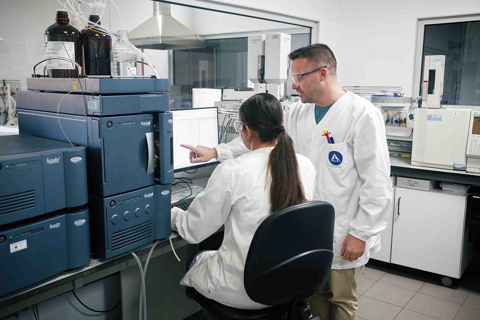 Two scientists in a laboratory wearing white lab coats and safety glasses, working together at a computer surrounded by liquid chromatography equipment and chemical bottles.