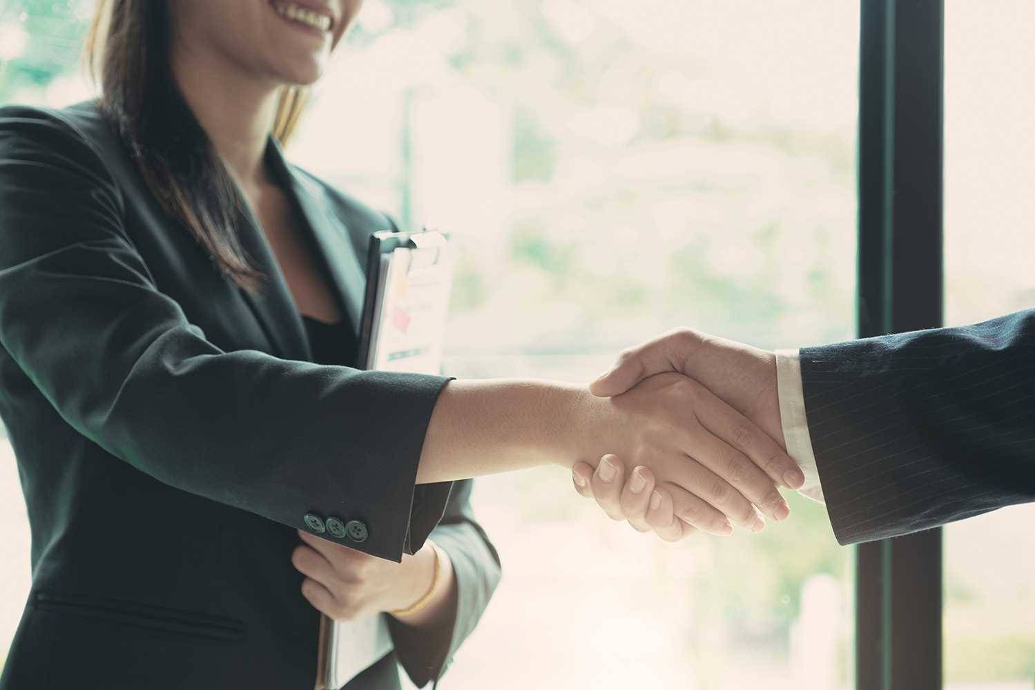 Dois profissionais em fato escuro a apertar as mãos, um deles com uma prancheta, num ambiente interior com luz natural.