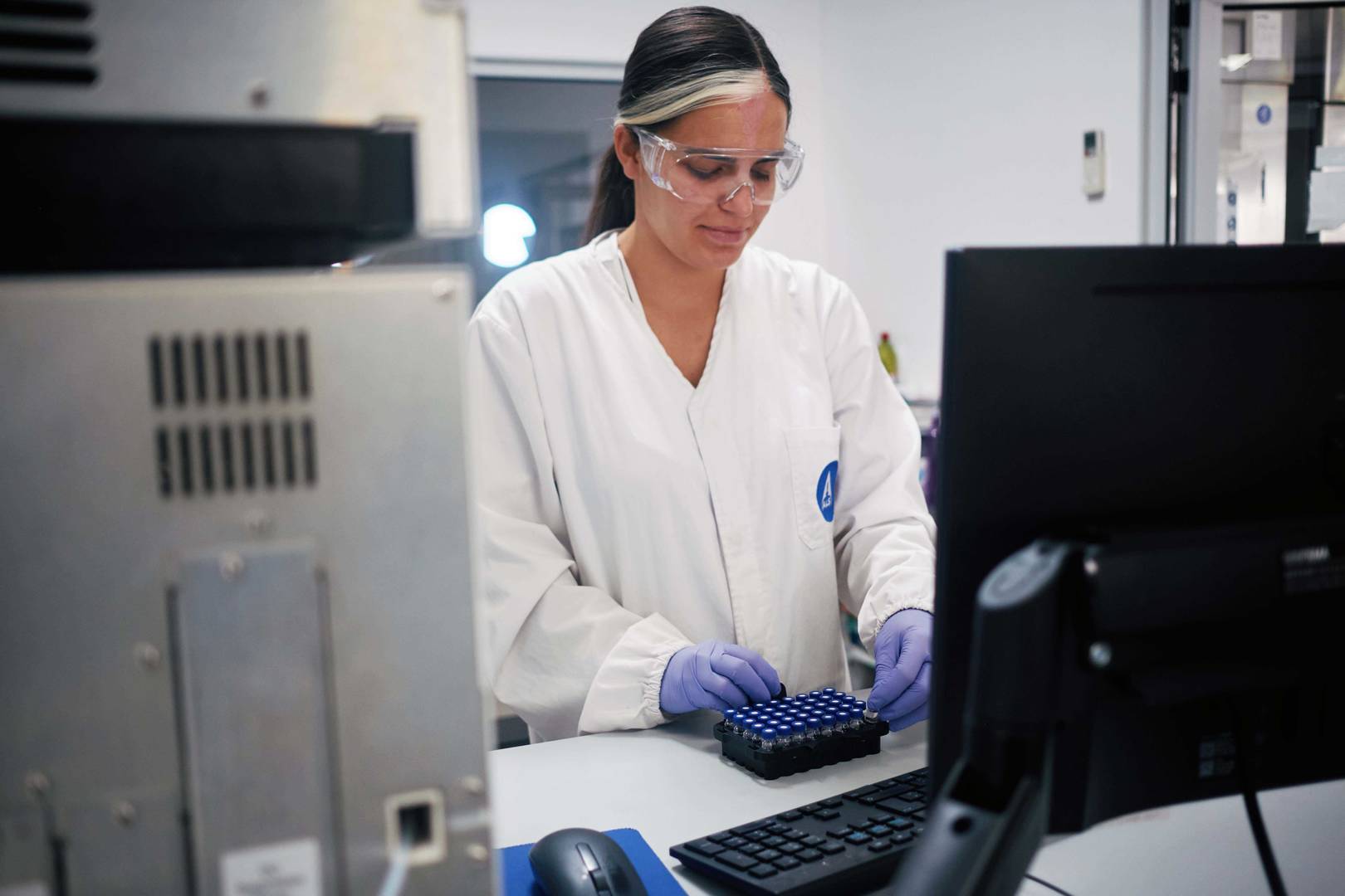 Pessoa com jaleco branco e luvas a trabalhar com bandeja de tubos de ensaio num laboratório com computador.