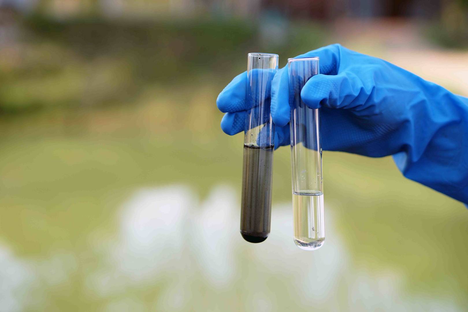 Mão com luva a segurar dois tubos de ensaio, um com líquido turvo e outro com líquido claro, representando contaminação.