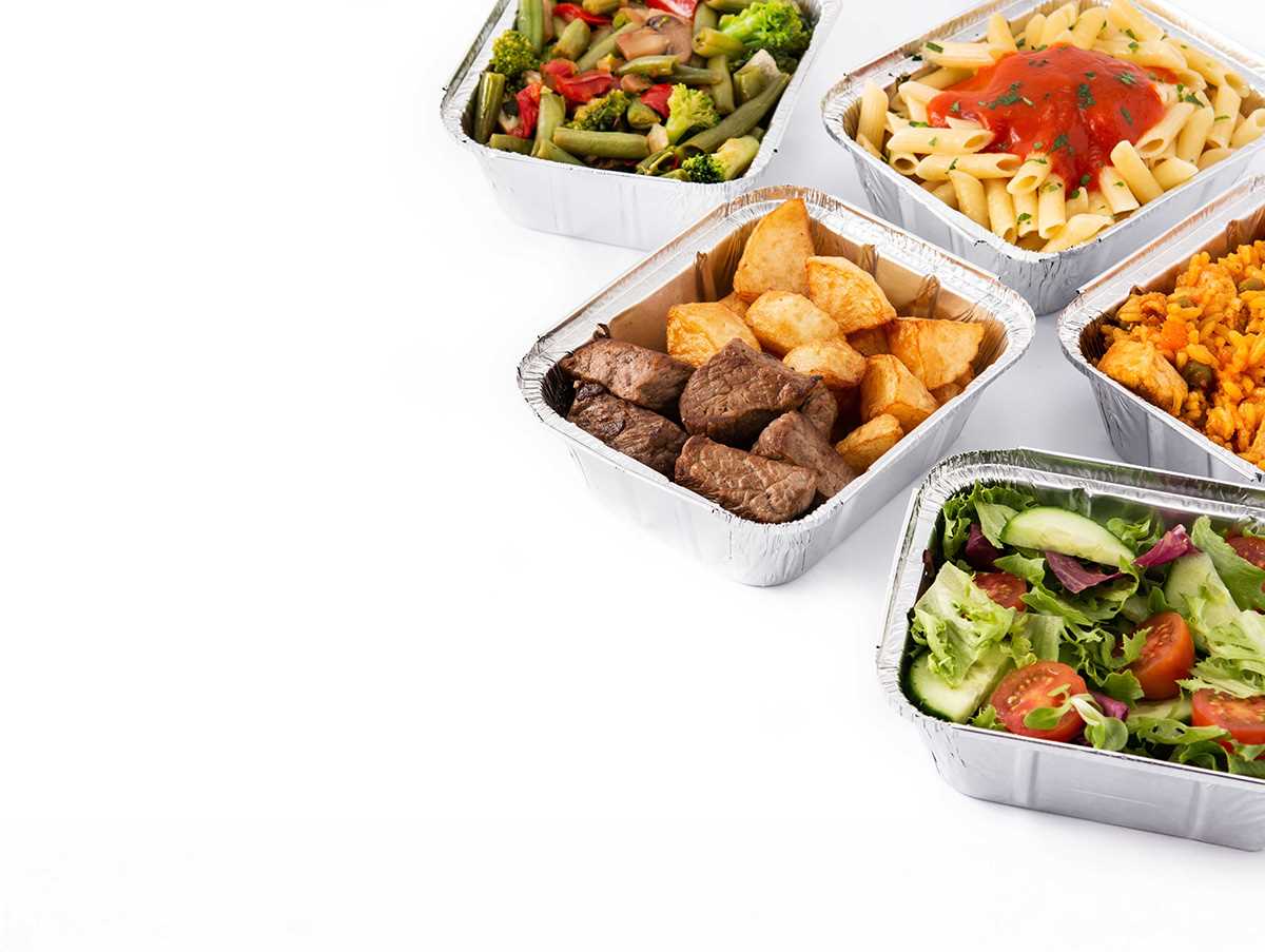 Recipientes de alumínio com diferentes refeições preparadas, incluindo salada com legumes frescos, massa com molho de tomate, legumes salteados, arroz e um prato com pedaços de carne e batatas.