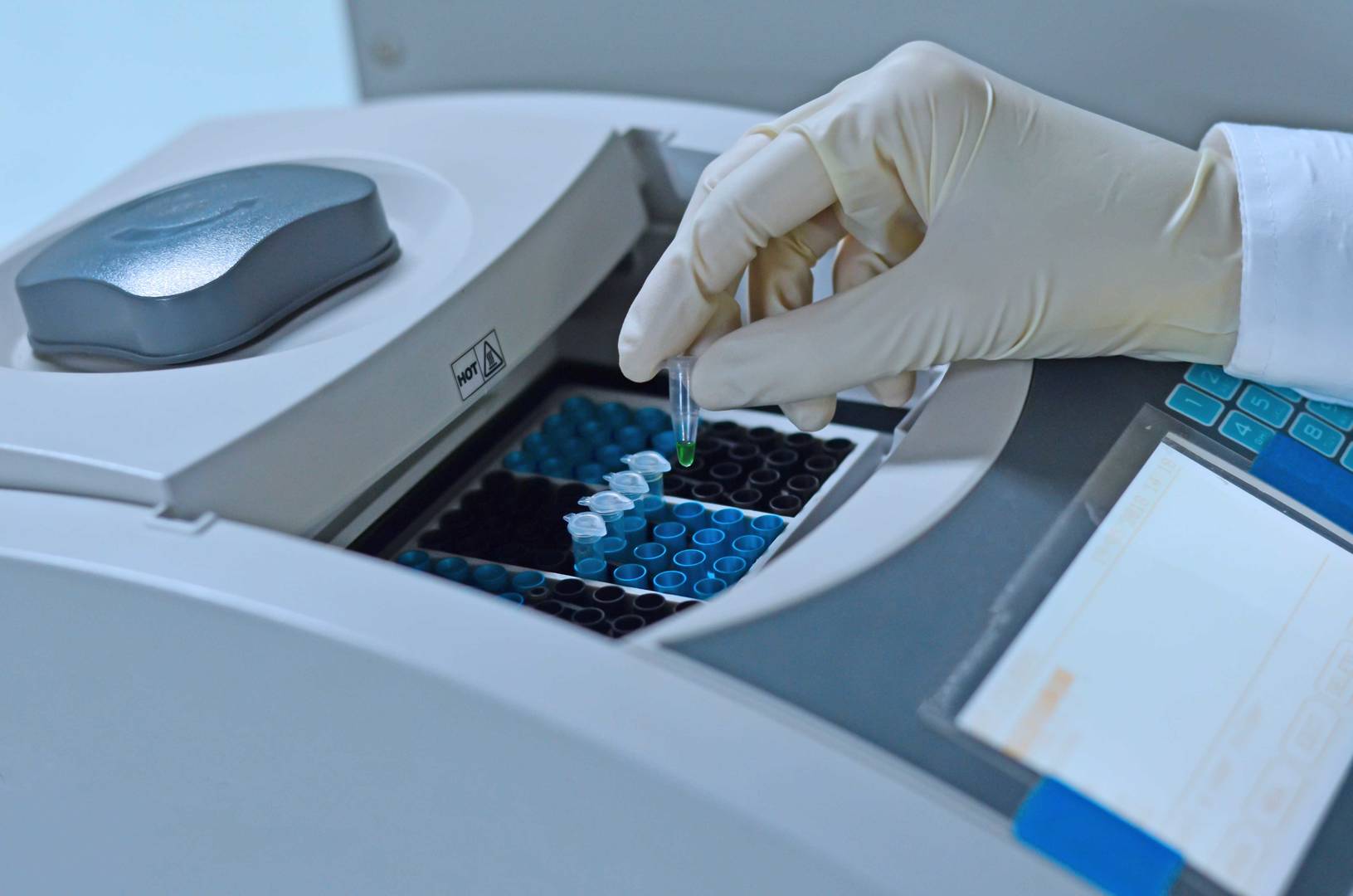 Mão com luva de látex a colocar um tubo de ensaio com líquido verde num equipamento laboratorial para análise, com vários tubos organizados em compartimentos.