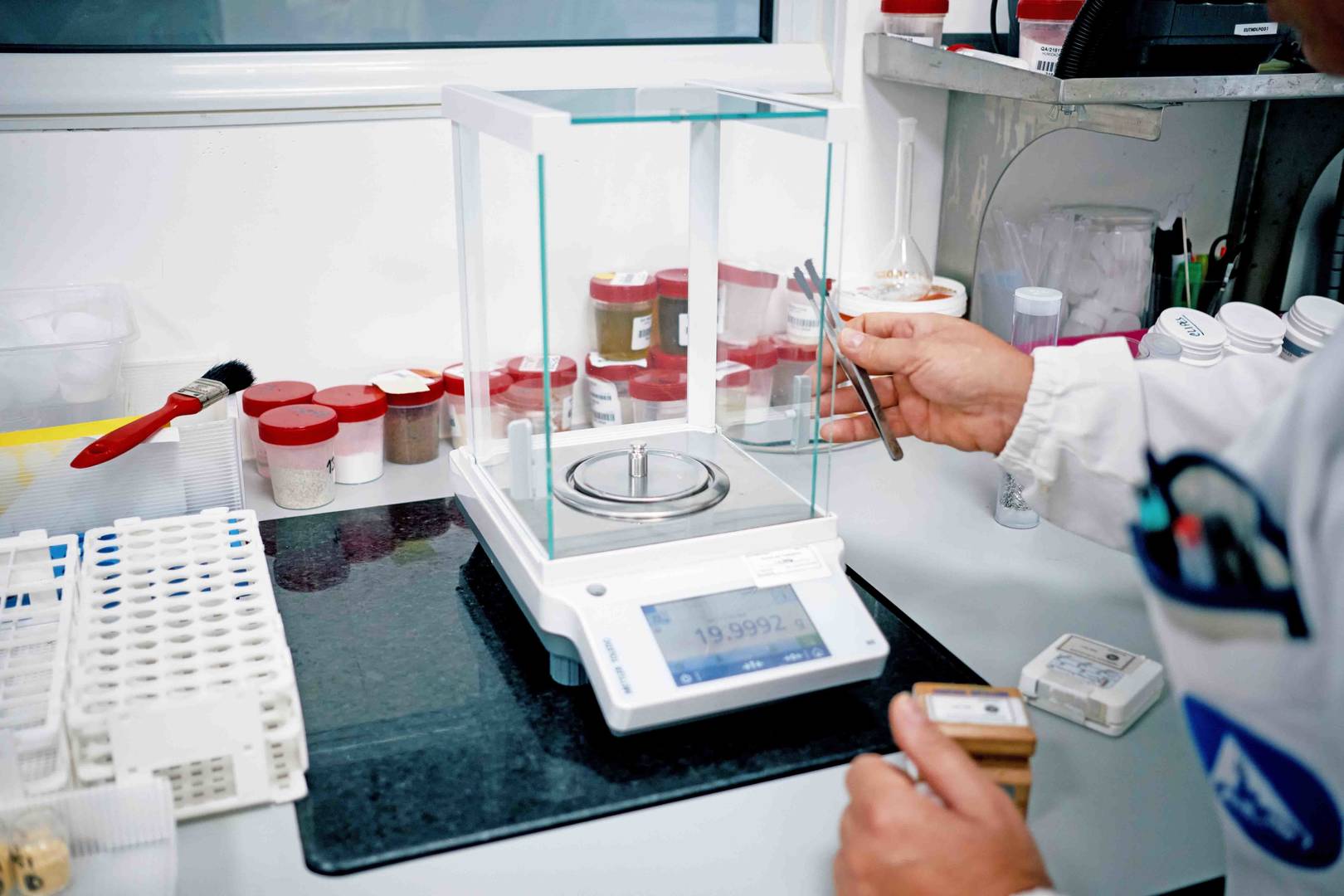 Pessoa utilizando uma balança analítica em um laboratório para pesar amostras com frascos ao fundo.