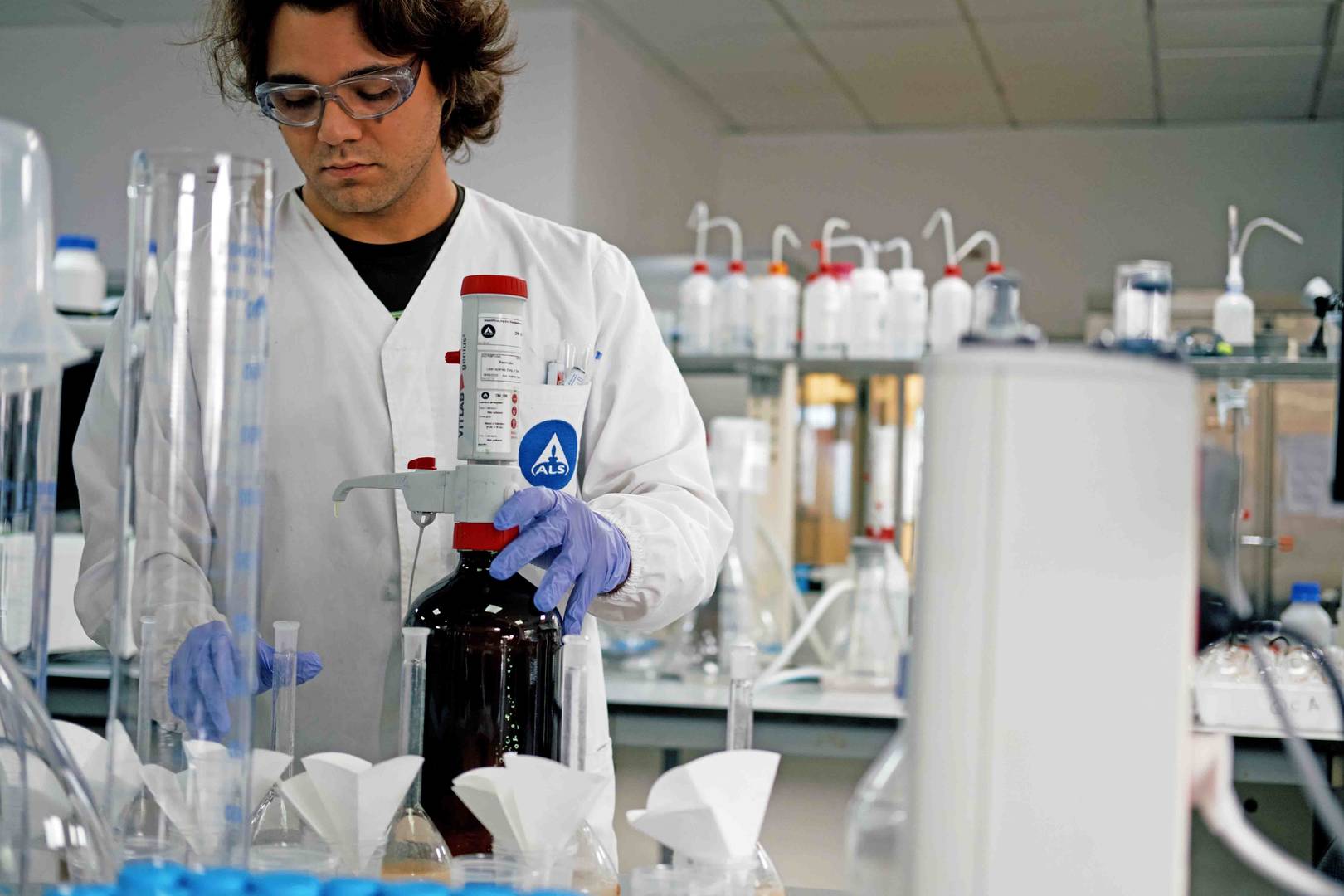 Pessoa com luvas e bata branca a utilizar frasco castanho com doseador num laboratório.