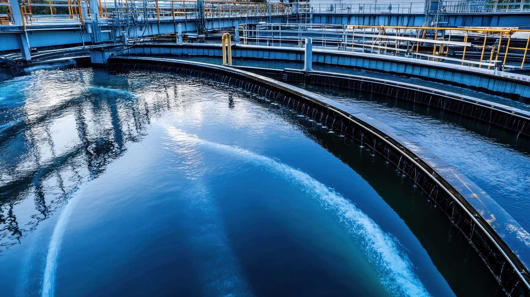 Vista de uma estação de tratamento de água com tanques circulares e estruturas metálicas ao redor. A água nos tanques está a ser movimentada por jatos, criando ondulações na superfície.