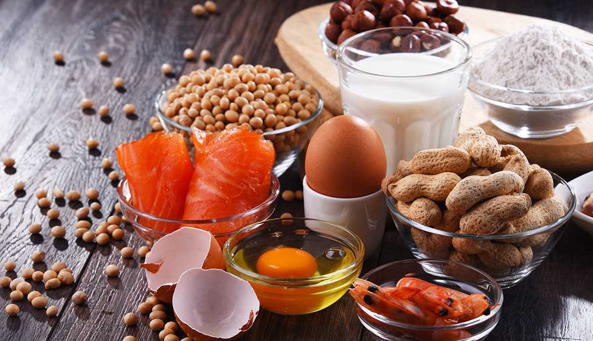 Composição de alimentos alergénicos comuns sobre uma mesa de madeira, incluindo ovos e cascas de ovo, peixe cru, camarão, leite, farinha de trigo, amendoins, frutos secos e soja, apresentados em tigelas e recipientes de vidro.