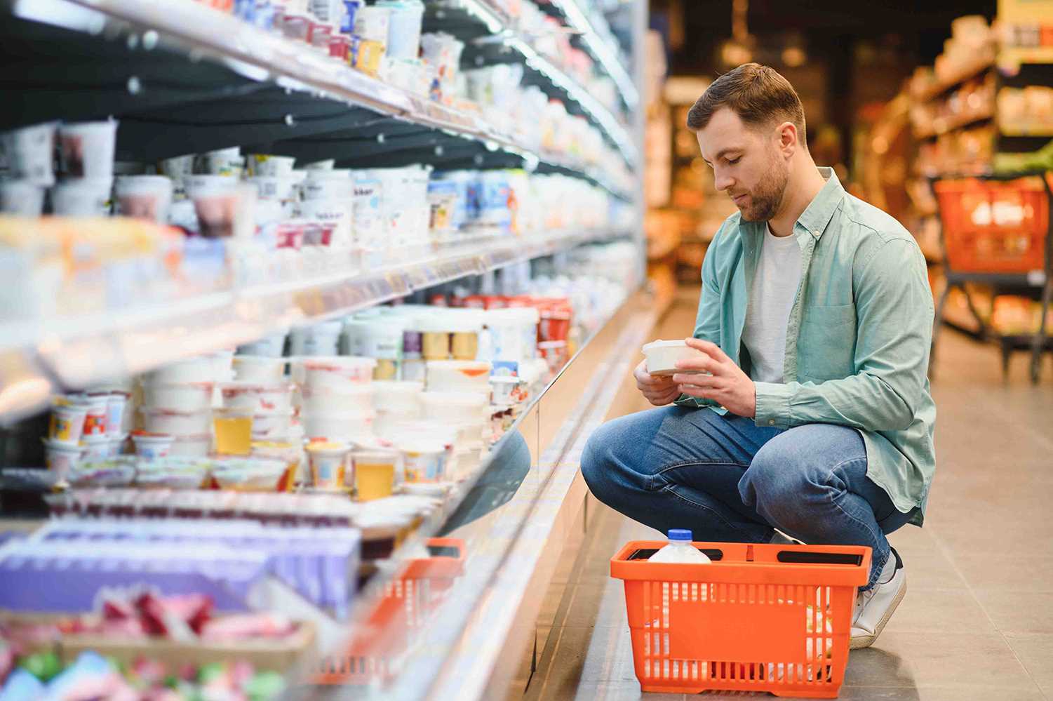 Homem ajoelhado no corredor de um supermercado, a examinar um produto junto a uma cesta de compras laranja.