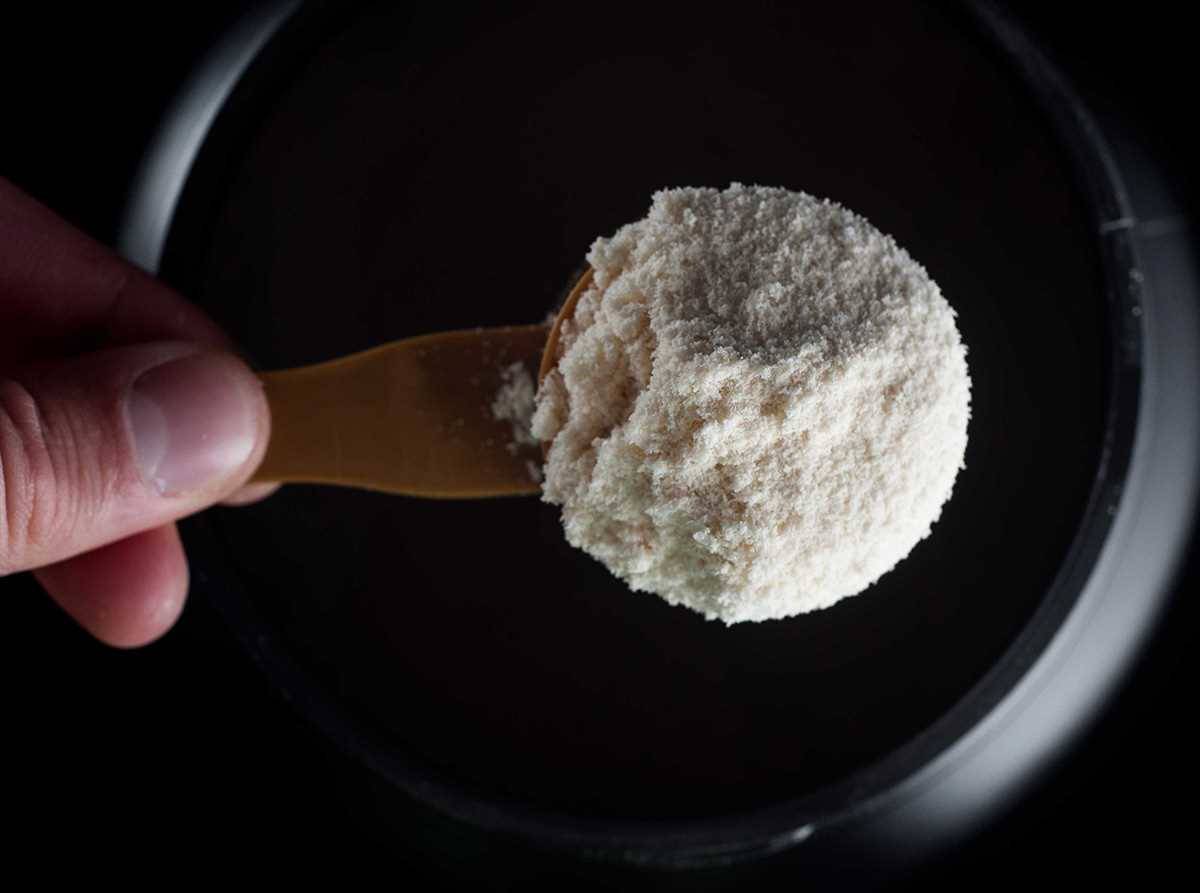 Pote de creatina em pó ao lado de uma colher medidora cheia, com o pó branco espalhado levemente sobre a mesa.