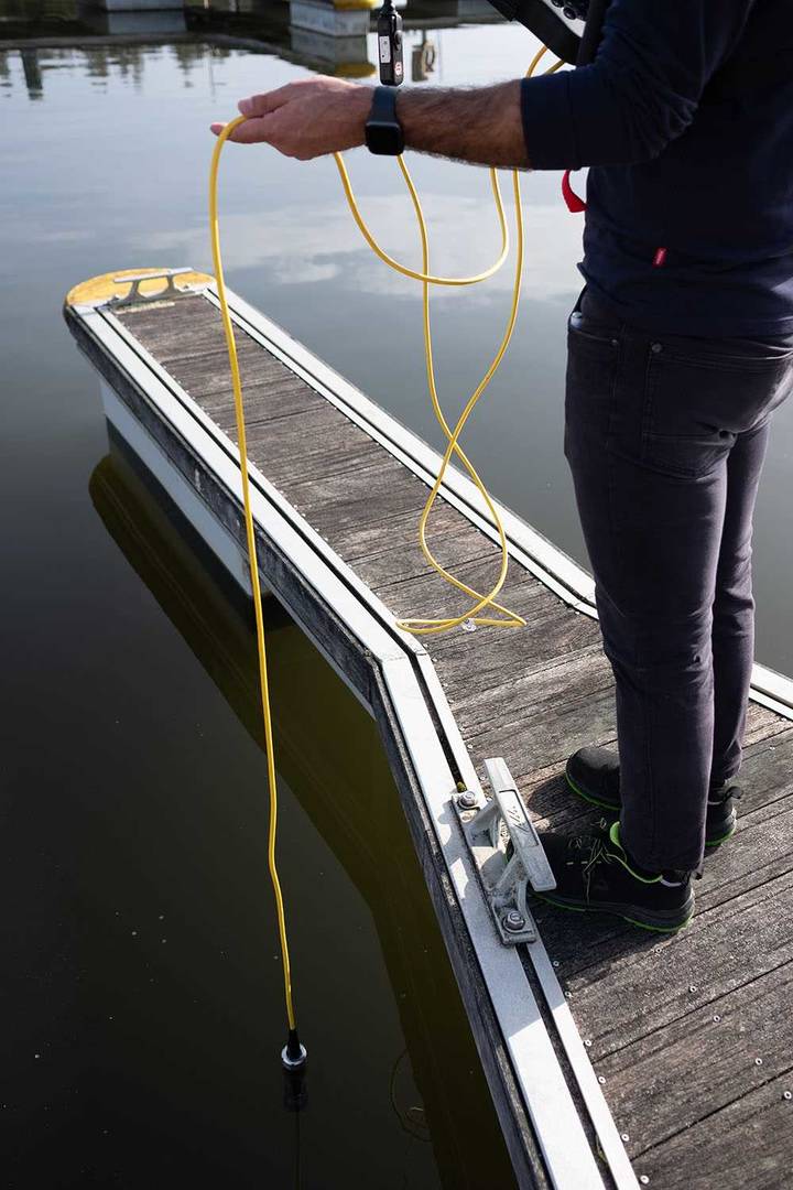 Técnico a efetuar medição da qualidade da água utilizando uma sonda com cabo amarelo a partir de um cais flutuante.