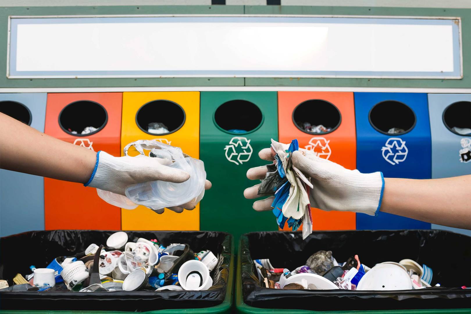 Duas mãos com luvas a separar resíduos plásticos para reciclagem, com contentores coloridos ao fundo identificados para diferentes tipos de materiais recicláveis.