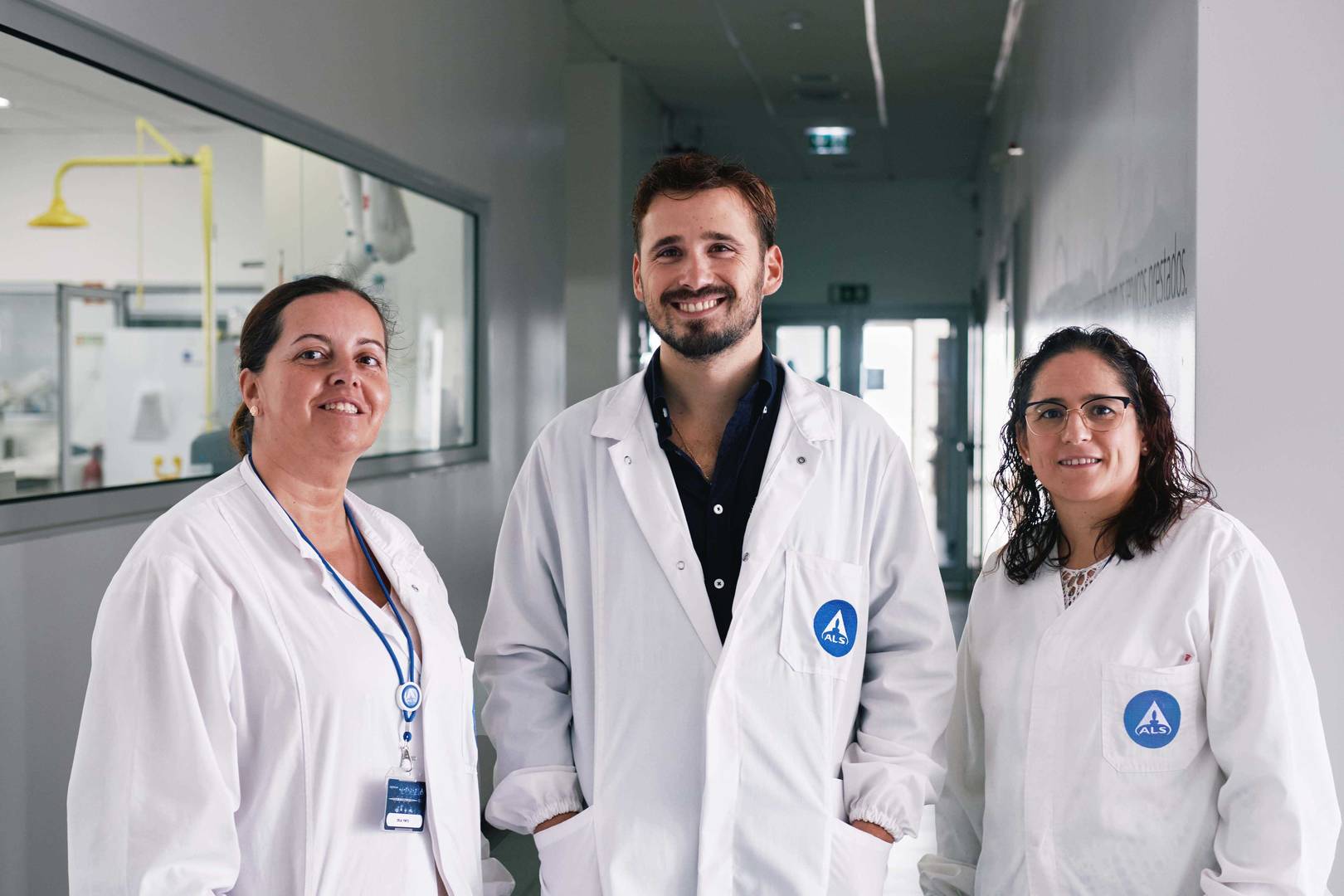 Três pessoas com batas brancas de laboratório a posar num corredor, com uma janela à esquerda mostrando parte de um espaço técnico.