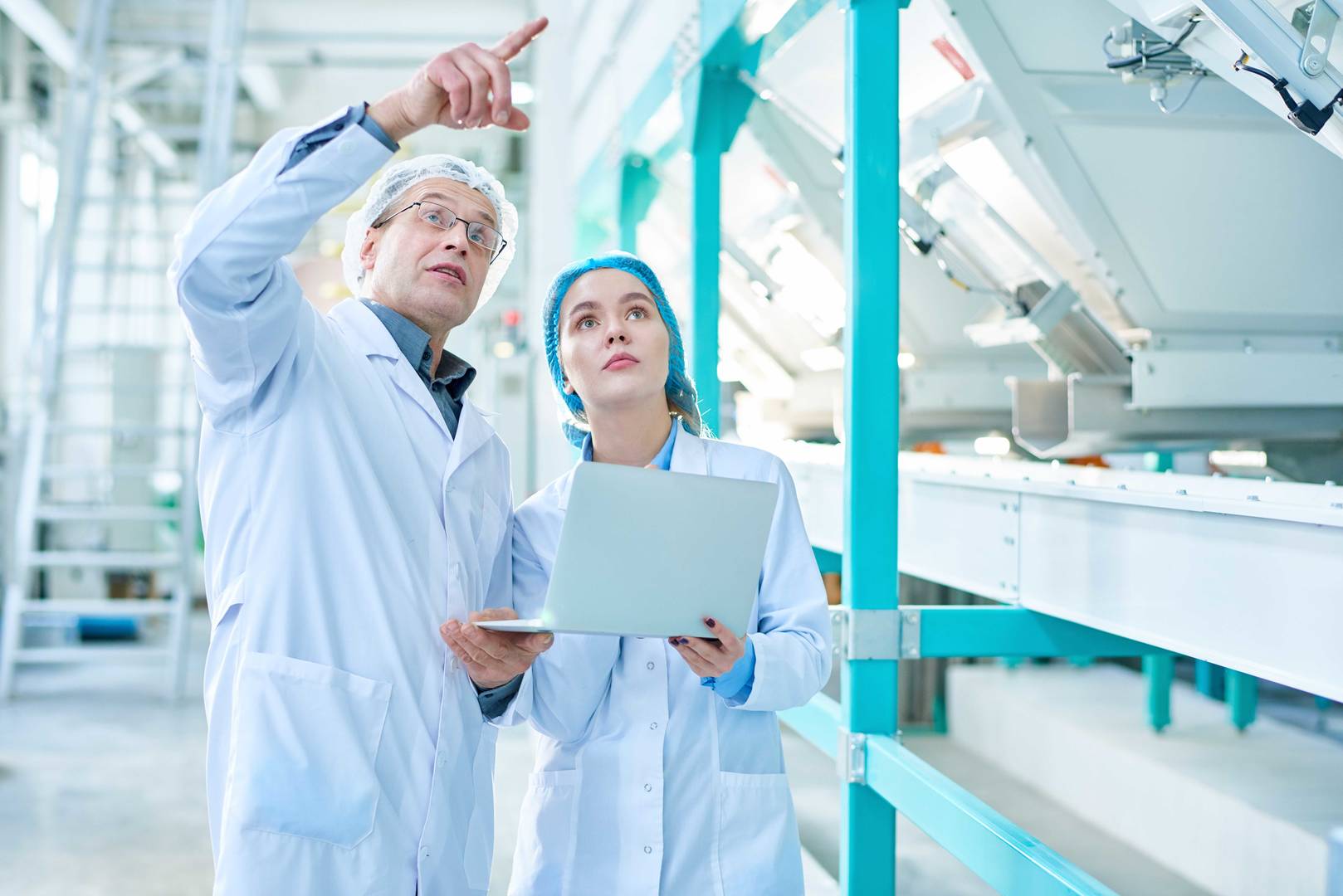 Portrét staršího továrního dělníka v laboratorním plášti, který vysvětluje pravidla stážistce a ukazuje vzhůru, zatímco stojí v čisté výrobní dílně moderního průmyslu.