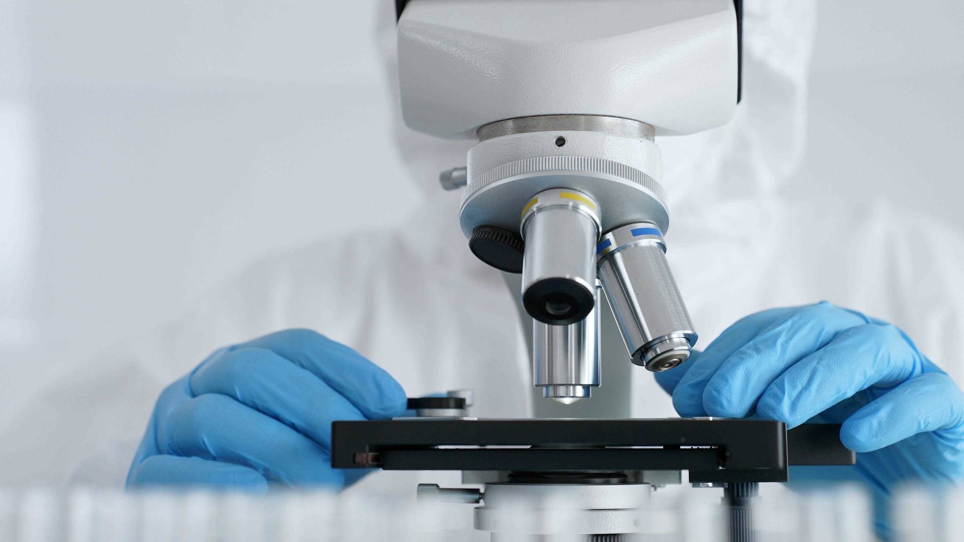 Vědecký výzkumník nebo virolog v ochranném oděvu nastavuje čočky mikroskopu naproti laboratorním zkumavkám s modrou kapalinou v laboratorním prostředí. Koncept medicíny, zdravotní péče a vědy.