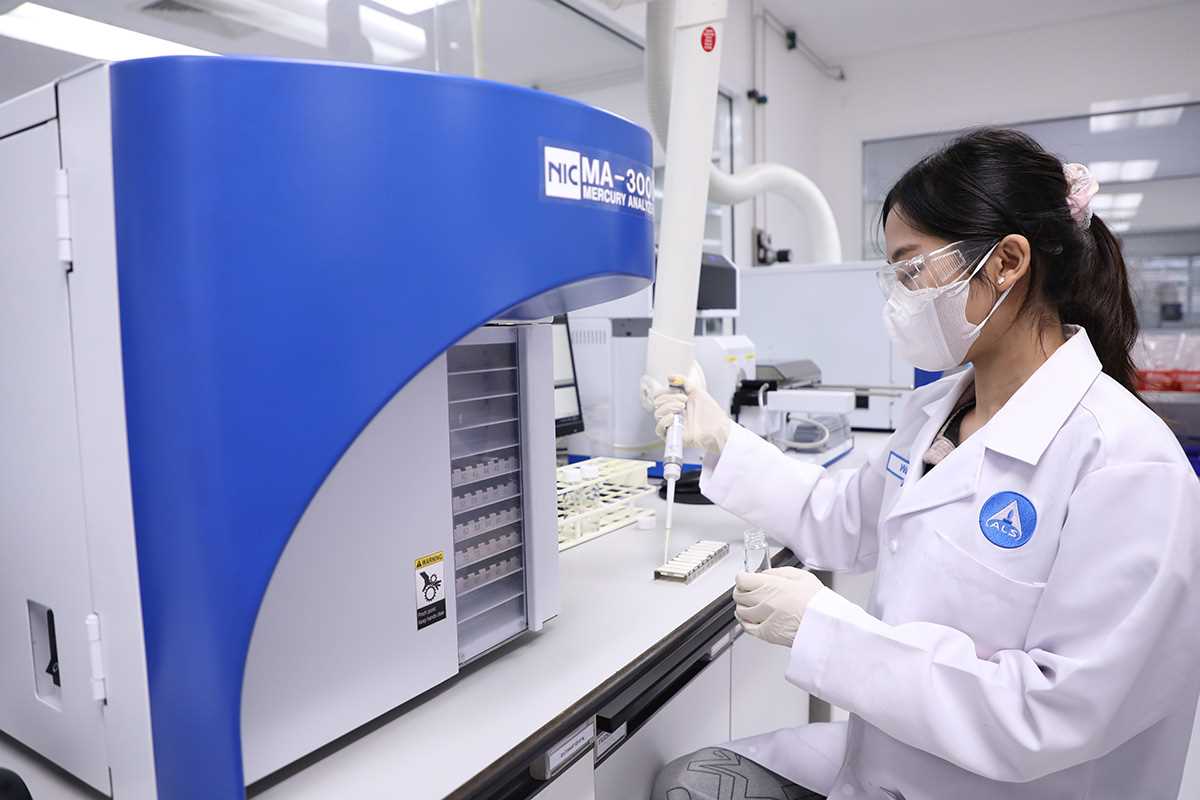 A person wearing a laboratory coat uses a pipette to handle samples next to a large blue analytical instrument inside a laboratory equipped with scientific workstations and devices.