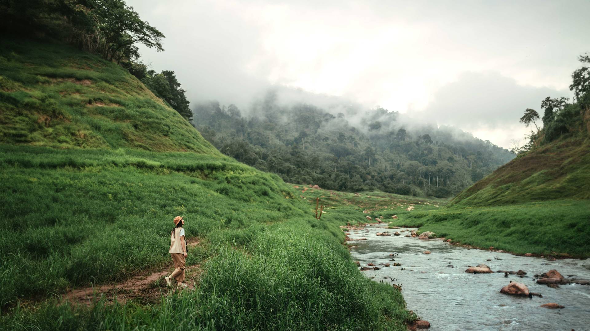 A person walks along a narrow dirt path beside a flowing river in a lush green valley, surrounded by hills covered in dense vegetation under an overcast sky.