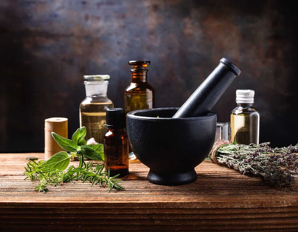 Herbal medicine setup on a wooden table, featuring a black mortar and pestle surrounded by fresh herbs, dried plants, and various small glass bottles filled with oils or extracts.