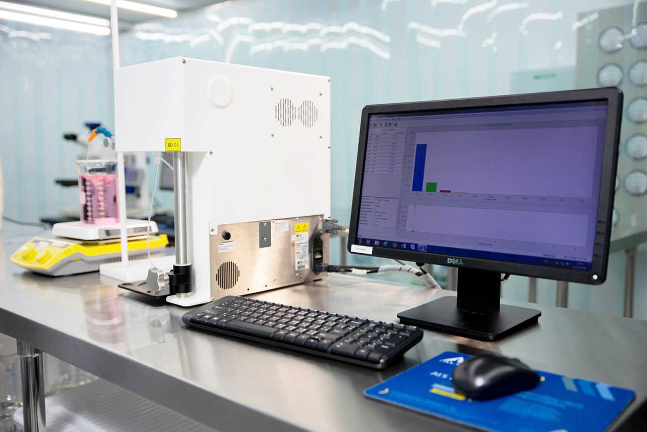 A liquid‑borne particle counting instrument connected to a computer displays analysis results on a laboratory workbench, with a beaker of pink liquid visible in the background.