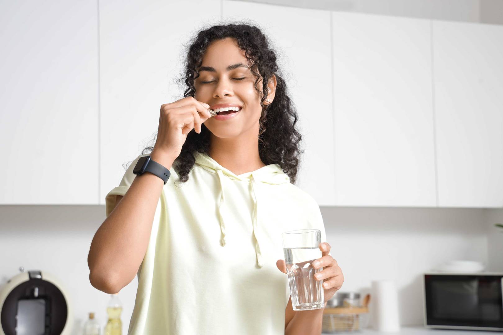 Bir mutfakta, bir eliyle yüzünü kapatırken diğer eliyle su bardağı tutan kişi.