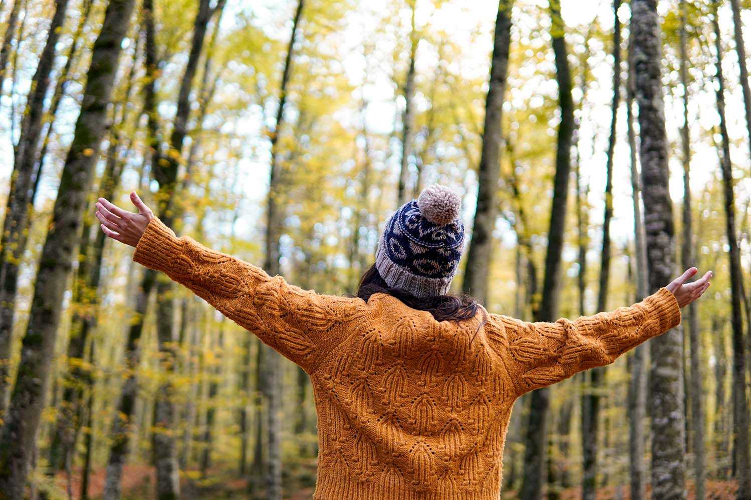 A person wearing an orange knitted sweater and a patterned winter hat stands with arms outstretched in a forest of tall trees with yellow autumn leaves.