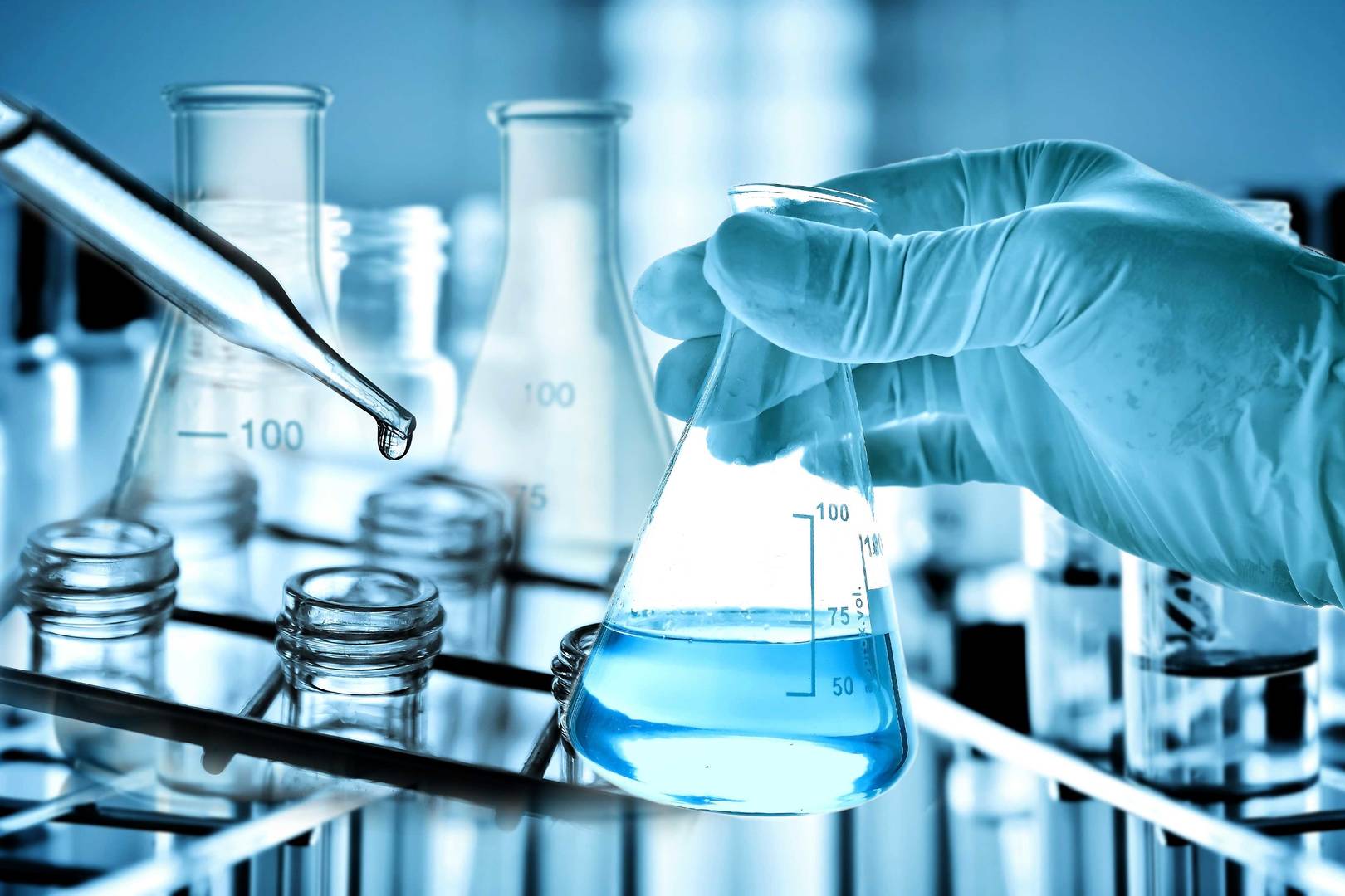 Laboratoriearbete med fysikalisk och kemisk analys – hand med blå handske håller Erlenmeyer-kolv med blå vätska bredvid provrör och pipett.