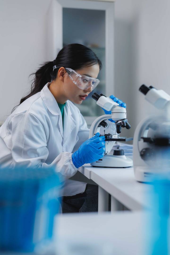 Laboratoriearbete med mikroskop, person i vit rock och blå handskar analyserar prover i forskningsmiljö.