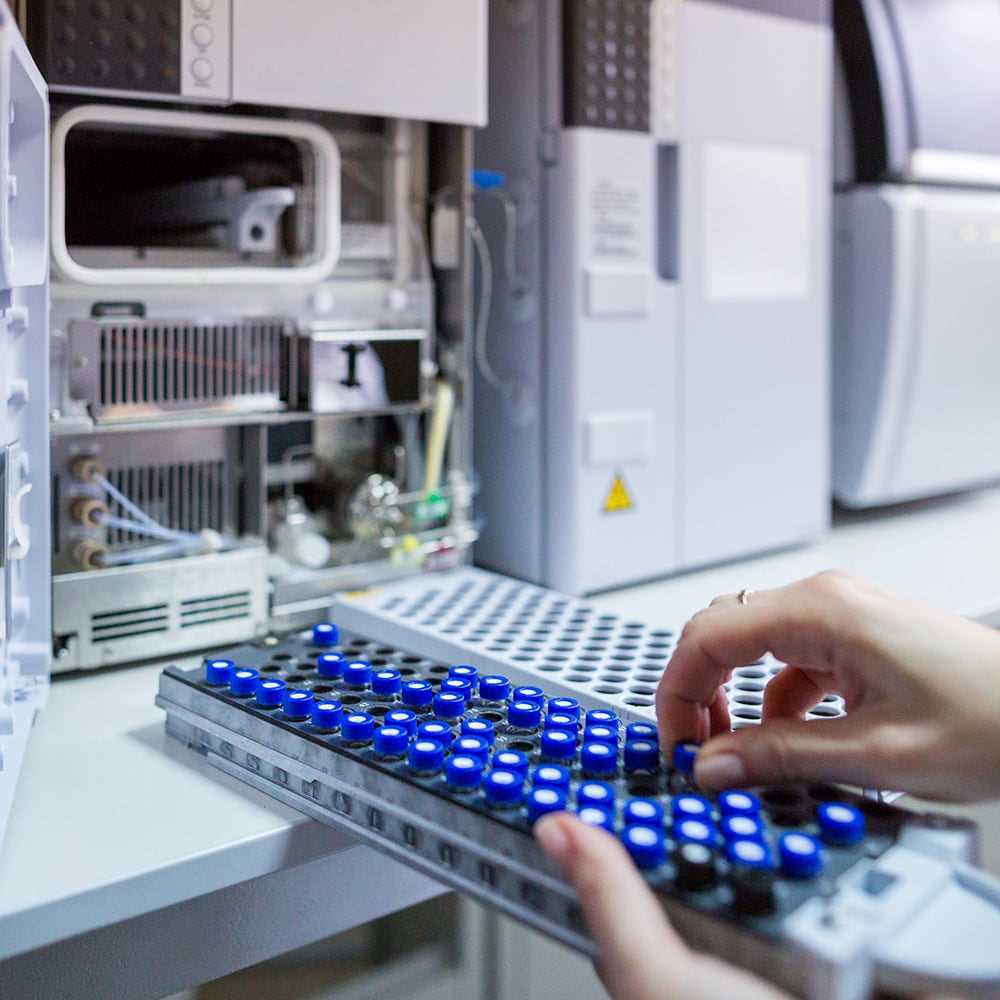 En person placerar en bricka med blå kapsylförsedda provrör i en avancerad analysinstrumentpanel i ett laboratorium