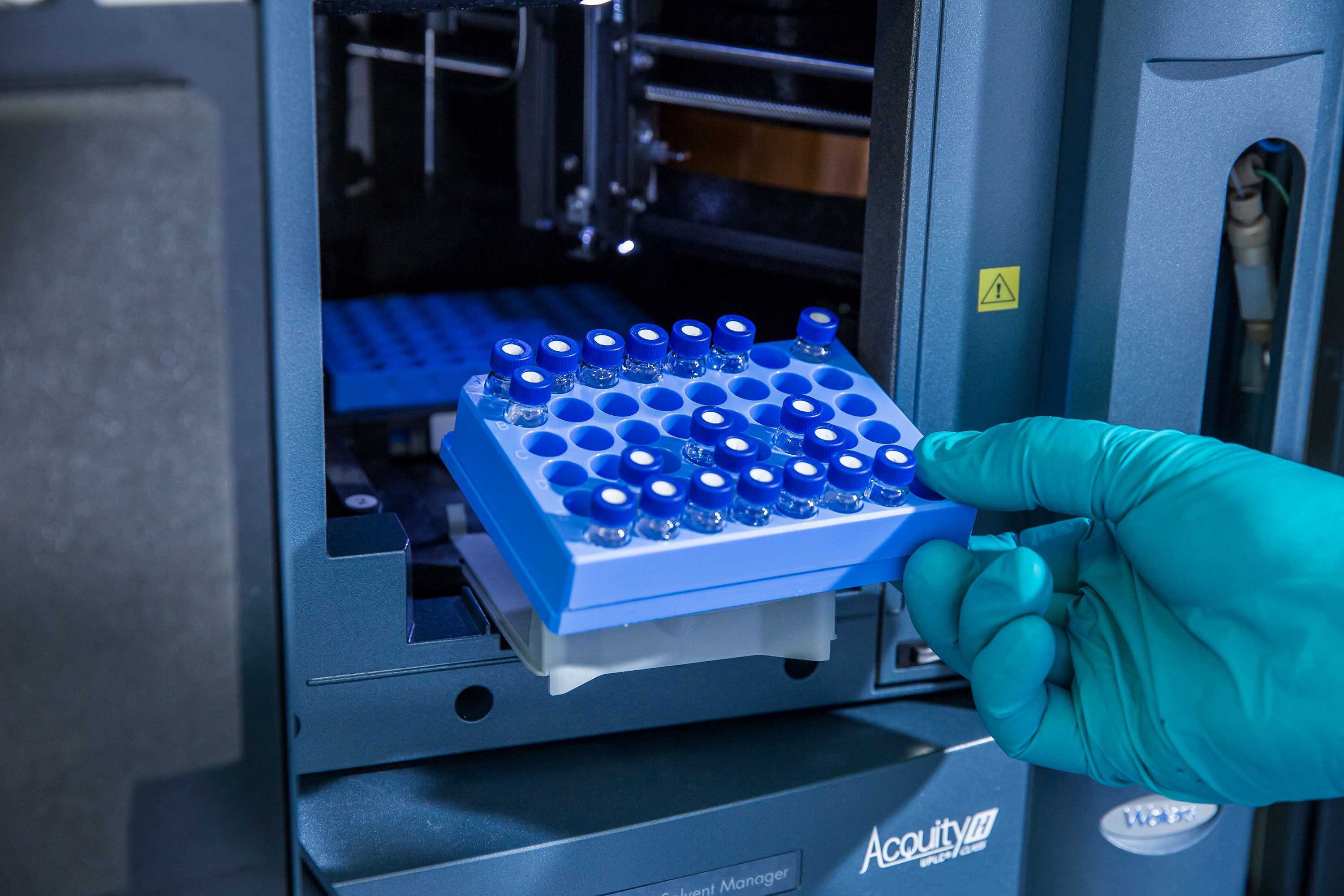 A gloved hand places a tray of small vials with blue caps into a laboratory instrument