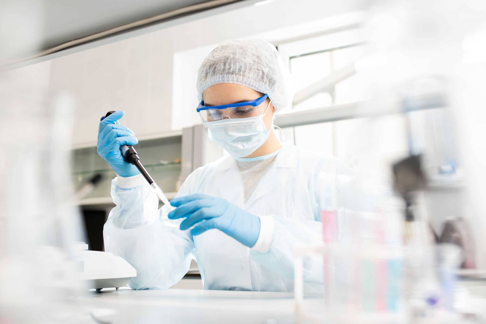 Microbiologist in sterile clothing and safety goggles adding reagent to a petri dish in a laboratory