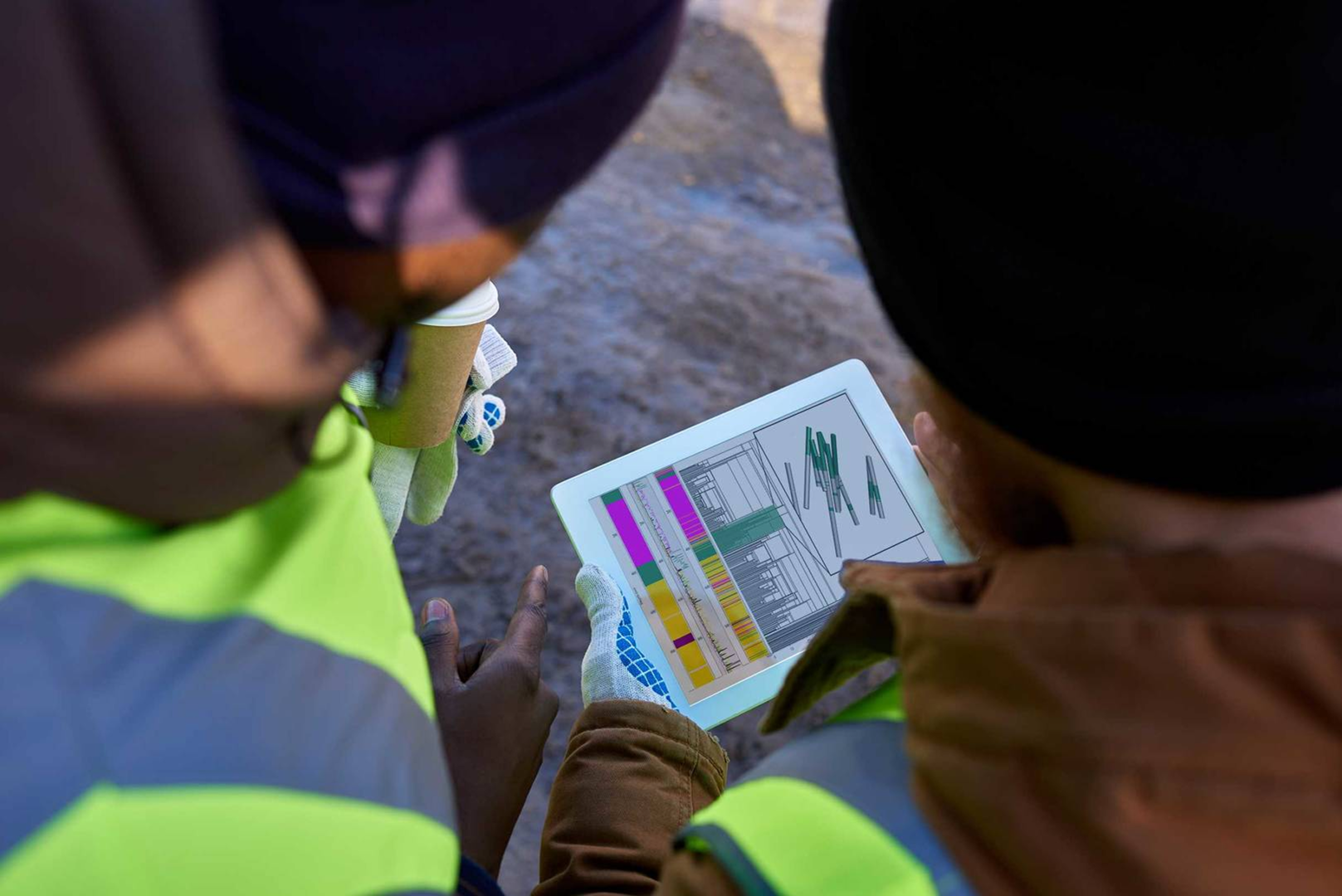 Overhead view of two field workers in high-visibility vests and beanies reviewing a tablet displaying colourful geological drill core logs, one holding a takeaway coffee cup, standing on a rough ground surface.